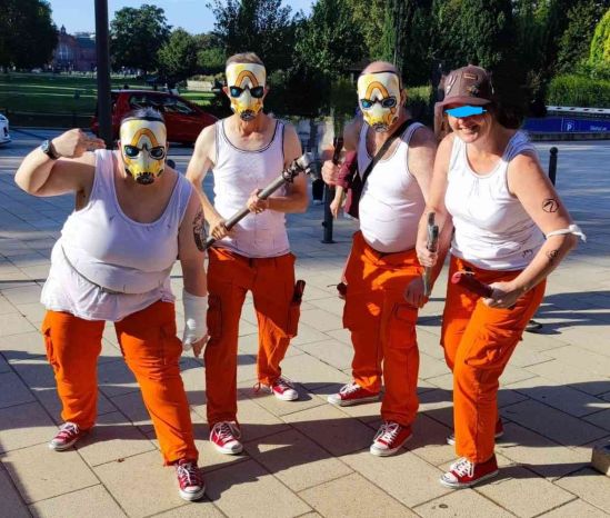 Vier Psychos von Borderlands in orangenen Hosen, weißen Unterhemden und Psychomasken, teilweise bewaffnet mit TNT-Stangen und verschiedenem Werkzeug, posieren für das Foto