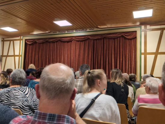 In einer Fachwerkwand befindet sich ein großer rötlich-brauner Vorhang. Davor sind die Stühle bereits mit Zuschauern besetzt - und über dem Vorhang steht in der Mitte - Fidelia Verein Ramsthal.
