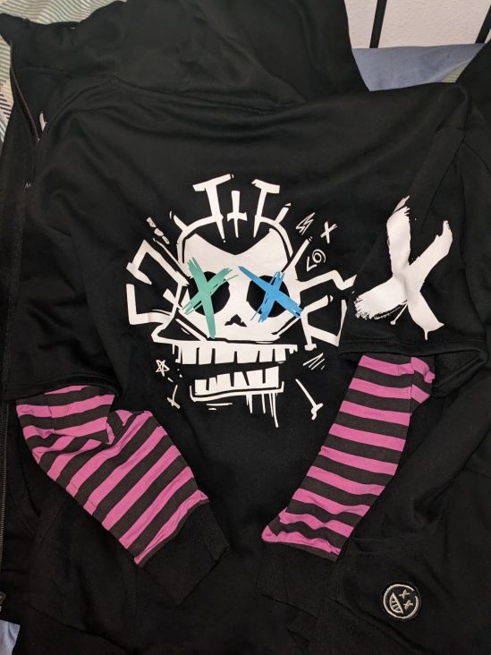Rückseite einer Jinx-Kapuzenjacke Grundfarbe ist Schwarz, auf der Rückseite ein großer Affentotenkopf im Jinx-Style Die Ärmel sind lila-scharz gestreift, rechts oben ist ein großes weißes Kreuz, unten ein Smiley.
