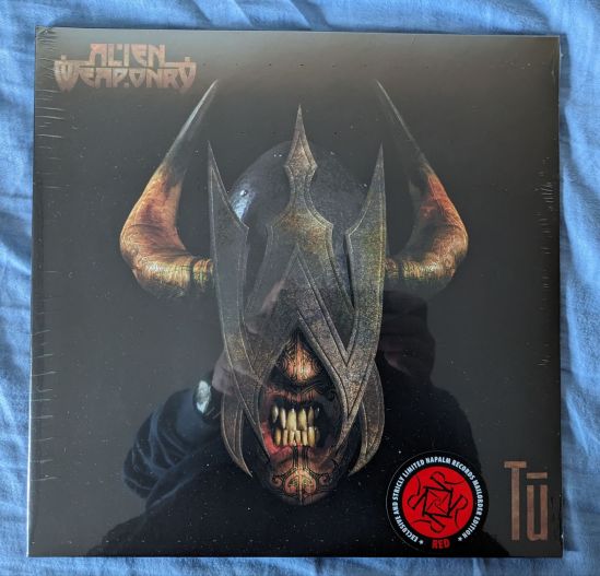Cover des Albums Tū von der Band Alien Weaponry Ein Kopf, verdeckt durch eine stilisierte Eisenmaske, mit gefletschten Zähnen und zwei Hörnern an beiden Seiten. Am Kinn unter den Zähnen und auf der Nasenspitze über den Zähnen sind Tatoos der Maori zu erkennen.