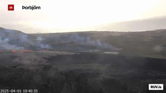 Bildschirmfoto der Eruption vom 01.04.2025 Livestream von RÚV. Unten links steht 2025-04-01 10:40:35 Oben links im Eck steht die Standortangabe der Kamera: Þorbjörn Im Hintergrund ist das sonnenbeschienene Meer zu sehen. Auf dem Land davor zieht sich eine Feuerlinie in der Mitte des Bildes bis etwa zur Mitte. Vor dieser Feuerlinie ist die sich ausbreitende Lava-Fläche zu erkennen. Von links nach rechts zieht sich der Verteidigungswall von Grindavík, der die Feuerlinie zu unterbrechen scheint, allerdings ist auf beiden Seiten des Walls Feuer zu sehen. Etwas weiter links auf dem grünen Feld ist eine weitere Rauchwolke zu erkennen, unter denen sich ebenfalls Flammen eines Spaltenausbriuchs befinden. Unten rechts steht RUV.is