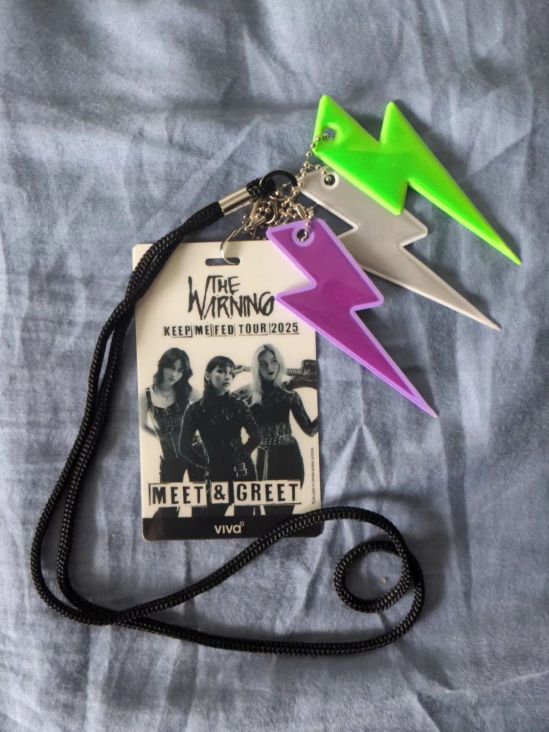 VIP-Anhänger für das Meet & Greet von The Warning Auf dem Anhänger ist ein Bild der drei Damen zu sehen. Der Name der Band (THE WARNING) und KEEP ME FED TOUR 2025 ist über ihnen geschrieben. Unten ist der Schriftzug MEET & GREET zu sehen. Am Anhänger sind drei stilisierte Blitzanhänger von einem Fan angebracht, in den Farben Lila, Weiß und Grün.