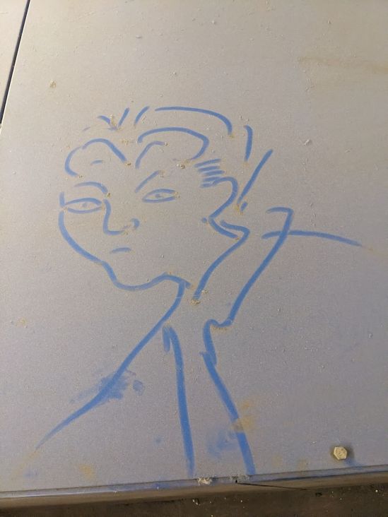 Zeichnung eines Gesichtes im Staub einer Abdeckung. Der Staub ist dreckig-gelb, wo das Gesicht gezeichnet wurde, ist es blau.