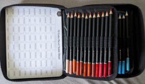 15 Buntstifte im Aufbewahrungs-Hefter. Links im Deckel ist ein Blatt für die 72 Farben des Buntstift-Hefters zum Testen, rechts 12 Stifte von hellorange bis rot, darunter noch 3 Stifte in hellblau