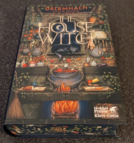 Cover des Buches "THE HOUSE WITCH" von Enelie Nikota von der Hobbit Presse / Klett-Cotta Das Titelblatt ist eine gemalte Küche mit gefülltem Topf über dem Feuer , Schneitbrettern links und rechts neben dem Topf, Gemüseschnippsel, die über dem Topf schweben und einer schwarzen Katze, die vor dem Fenster liegt. Die Seiten haben einen Farbschnitt.