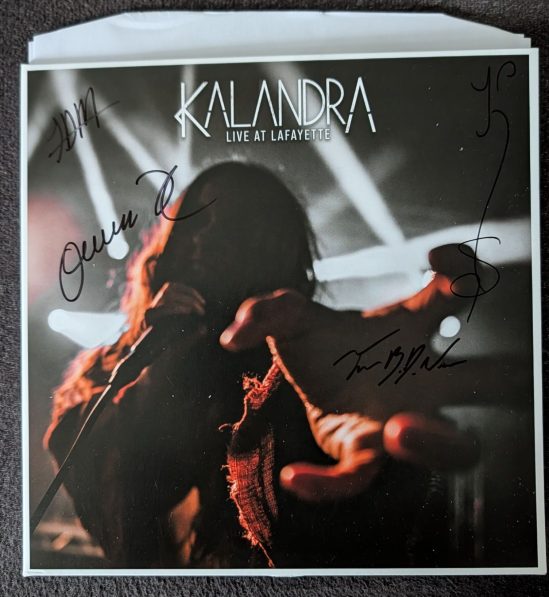 Cover des Albums KALANDRA LIVE AT LAFAYETTE Katrine greift singend Richtung Kamera, das Gesicht verdunkelt, im Hintergrund Lichtstrahlen Das Cover ist signiert.